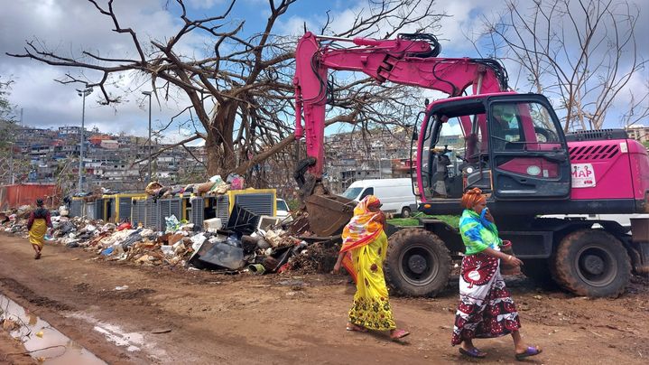 Dos mujeres pasan delante de una máquina que limpia un montón de residuos, en Koungou, al norte de Mayotte, el 27 de diciembre. (AGATHE MAHUET / FRANCIA INFORMACIÓN / RADIO FRANCIA)