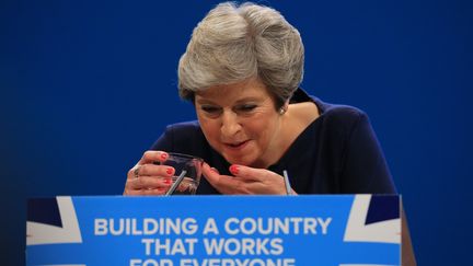 Theresa May, en pleine quinte de toux, boit de l'eau au cours de son discours devant les conservateurs britanniques, mercredi 4 octobre 2017 à Manchester. (LINDSEY PARNABY / ANADOLU AGENCY / AFP)