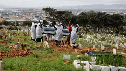 Dans un cimetière de Cape Town, sur la côte sud-ouest d'Afrique du Sud, des employés des pompes funèbres, masqués et revêtus de blouses, portent le cercueil d'un homme de 51 ans, mort du Covid-19 (photo prise le 12 mai 2020). (SUMAYA HISHAM / REUTERS)