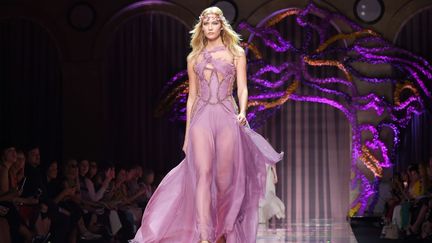 Un tapis de 25.000 orchidées jaunes et mauves -protégées par des plaques de verre- en guise de podium. Le ton est donné : tout en transparences et avec une abondance de volants, le flower power imprègne la collection Atelier Versace. De fines couronnes sont portées sur des chevelures détachées et des chaussures à plateformes évoquent les années 70'. La femme Versace habituellement conquérante et sexy cède la place, cette saison, à une version plus douce. Dans sa garde-robe : des robes délicates faites de couches de mousseline de soie transparente, aux manches évasées pour certains modèles, des robes, asymétriques, à la traîne volumineuse, avec des bords qui s'effilochent pour donner un effet éthéré à la silhouette. Des fleurs semblent flotter sur une longue robe bordée de velours, de minces sangles retiennent un chandail surdimensionné tandis que des bandes de mousseline de soie se mêlent aux cristaux Swarovski et à la cotte de mailles. La palette est délinée dans du gris colombe, du rose poudré, du vert fané et du lilas. Cette femme "fleurs" n'est cependant pas si sage qu'il n'y parait avec ses décollettés audacieux et ses savantes découpes ! 
