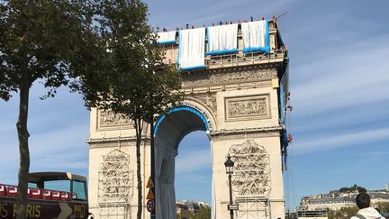 Dimanche, les&nbsp;badauds se regroupent du côté de la façade avenue des Champs Elysées alors que les premiers rouleaux de tissu commencent à être déroulés. Au premier plan, à l'entrée du souterrain menant au monument, on peut voir le projet tel que l'a imaginé Christo. (VALERIE GAGET / FRANCE TELEVISIONS)