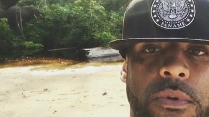 Booba et l'éléphant aux dangereuses défenses que l'on devine en arrière plan au milieu des arbres.
 (Instagram Booba Official)