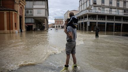 Un homme porte son fils et constate l'étendue des dégâts, à Lugo non loin de Ravenne le 18 mai 2023 après les inondations causées par des pluies massives sur la région nord-est d'Emilie-Romagne (FEDERICO SCOPPA / AFP)