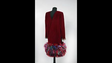 « BOUJOUR TRISTESSE ». Robe à effet portefeuille en velours de soie grenat, encolure en pointes, poignets zippés gansés d’un galon perlé et strassé tutti frutti, rappel à l’ourlet garni de plumes d’autruche lustrées à la couleur
 (DR)